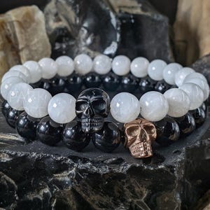 May include: Two beaded bracelets. One bracelet features white, round beads, and the other has black, round beads. Two skull charms, one black and one bronze, are placed between the bracelets. The bracelets are displayed on a dark, textured surface.
