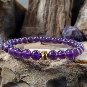 May include: A purple amethyst gemstone bracelet with a gold bead accent. The bracelet is on a textured rock surface.