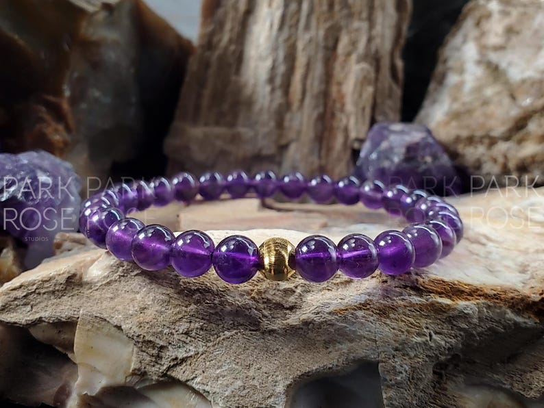 May include: A purple amethyst gemstone bracelet with a gold bead accent. The bracelet is on a textured rock surface.