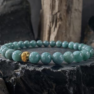 May include: A beaded bracelet featuring round, light teal beads with a single gold-colored bead. The bracelet is displayed on a dark, textured surface, with a natural wood element in the background. The beads are approximately 0.3 inches in diameter.