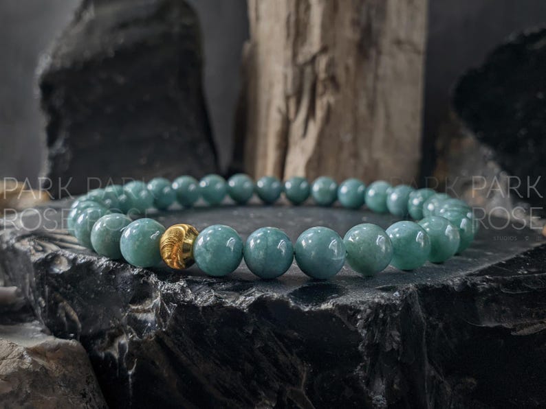 May include: A beaded bracelet featuring round, light teal beads with a single gold-colored bead. The bracelet is displayed on a dark, textured surface, with a natural wood element in the background. The beads are approximately 0.3 inches in diameter.