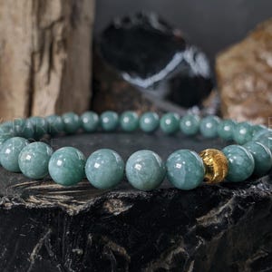 May include: A beaded bracelet featuring round, pale green jade stones. One gold-colored bead is incorporated into the design. The bracelet is displayed on a dark, textured surface with a blurred background of wood and stone.