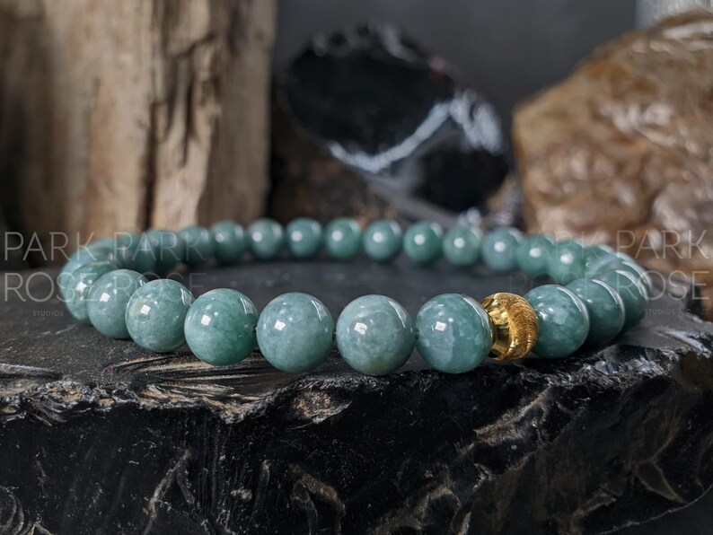 May include: A beaded bracelet featuring round, pale green jade stones. One gold-colored bead is incorporated into the design. The bracelet is displayed on a dark, textured surface with a blurred background of wood and stone.