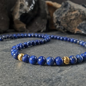 May include: A necklace featuring deep blue, spherical beads interspersed with gold-colored beads. The necklace is displayed on a dark, textured surface, with blurred dark rocks in the background. The necklace is a jewelry item.