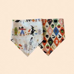 May include: Two triangular pet bandanas featuring Halloween motifs. The left bandana is off-white with illustrations of a scarecrow, a witch, a skeleton, and ghosts. The right bandana features a diamond pattern with icons of pumpkins, ghosts, and cats.