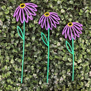 May include: Three purple metal flower garden stakes with yellow centers and green stems and leaves. The stakes are arranged in a row against a backdrop of green foliage. These decorative stakes add a touch of whimsy to any garden or outdoor space.
