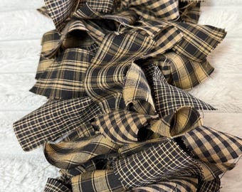 May include: Close-up of a decorative garland made of layered fabric strips. The fabric features a black and tan plaid pattern. The garland is arranged in a ruffled, layered design, creating a textured and visually appealing effect. The overall theme is rustic and farmhouse-style decor.