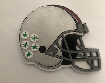 May include: A silver football helmet with a gray facemask and a red stripe on top. Six white circles with green leaf designs are on the side of the helmet. The helmet is a sports-themed item.