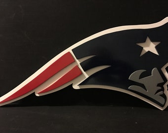 May include: A layered wooden sign displaying the New England Patriots logo. The design features a white star, a grey profile, and red and white elements set against a navy blue backdrop. This sign is a decorative item, suitable for display.