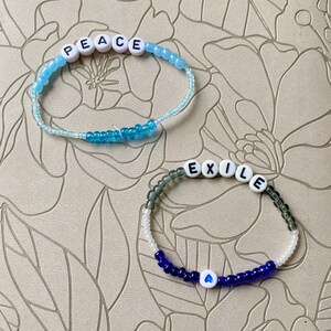 May include: Two beaded bracelets with white letter beads spelling out the words "PEACE" and "EXILE". The "PEACE" bracelet has blue and white beads, while the "EXILE" bracelet has gray, white, and blue beads. A blue heart bead is on the "EXILE" bracelet.