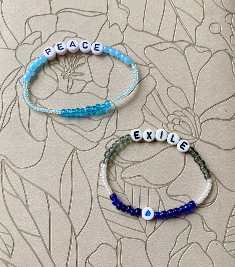 May include: Two beaded bracelets with white letter beads spelling out the words "PEACE" and "EXILE". The "PEACE" bracelet has blue and white beads, while the "EXILE" bracelet has gray, white, and blue beads. A blue heart bead is on the "EXILE" bracelet.
