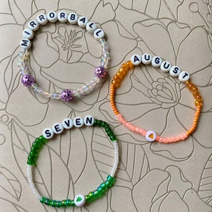 May include: Three beaded bracelets with white letter beads spelling out the words "MIRRORBALL", "AUGUST", and "SEVEN". Each bracelet has a different color scheme and a small heart charm.