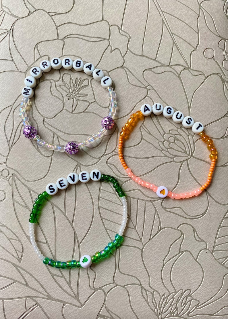 May include: Three beaded bracelets with white letter beads spelling out the words "MIRRORBALL", "AUGUST", and "SEVEN". Each bracelet has a different color scheme and a small heart charm.