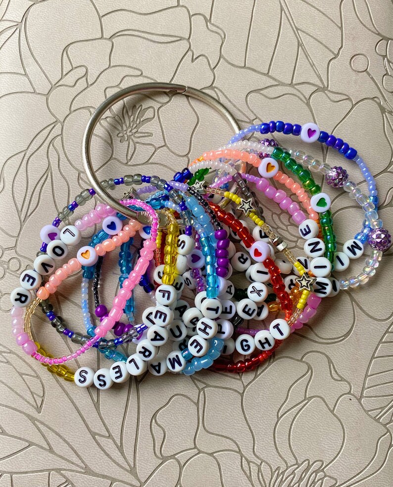 May include: A collection of colorful beaded bracelets with letter beads spelling out words like "FEARLESS", "LOVE", "NOW", and "BRIGHT".