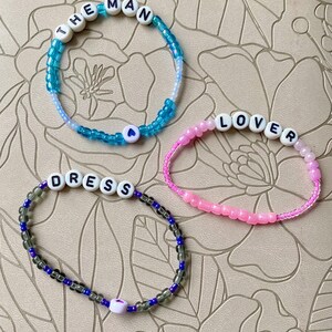 May include: Three beaded bracelets with the words "THE MAN", "LOVER", and "DRESS" spelled out in white beads. Each bracelet has a small heart charm.