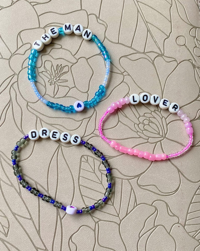 May include: Three beaded bracelets with the words "THE MAN", "LOVER", and "DRESS" spelled out in white beads. Each bracelet has a small heart charm.