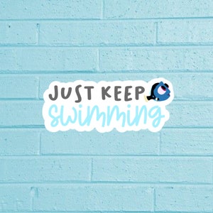 Può includere: Un adesivo blu e bianco con il testo "Just Keep Swimming" e un pesce blu dei cartoni animati con un occhio bianco.