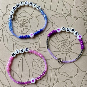 May include: Three beaded bracelets with the words "MIDNIGHTS", "OURS", and "MINE" spelled out in white beads. Each bracelet has a small heart charm and is made with a gradient of blue, purple, and pink beads.