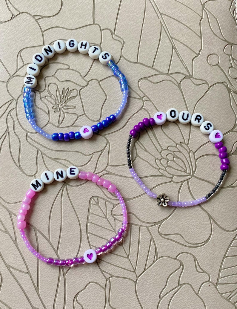 May include: Three beaded bracelets with the words "MIDNIGHTS", "OURS", and "MINE" spelled out in white beads. Each bracelet has a small heart charm and is made with a gradient of blue, purple, and pink beads.