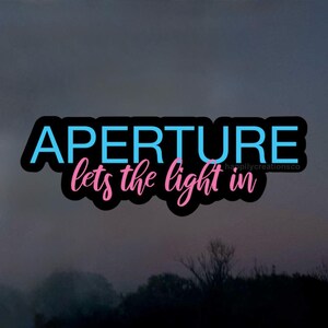 May include: A sticker featuring the word "APERTURE" in light blue, above the words "lets the light in" in pink, all with a black outline. The background is a blurred, dark landscape with a tree line.