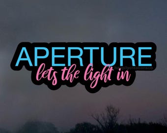 May include: A sticker featuring the word "APERTURE" in light blue, above the words "lets the light in" in pink, all with a black outline. The background is a blurred, dark landscape with a tree line.