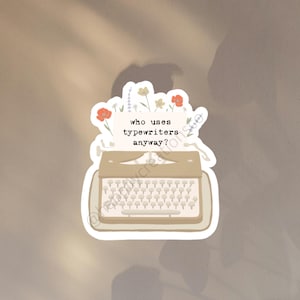 May include: A sticker with a vintage typewriter design and the phrase "who uses typewriters anyway?" in a playful font. The design incorporates floral elements in red, yellow, and purple, set against a neutral backdrop.