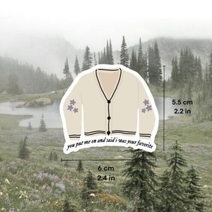 May include: A beige cardigan with black trim and three purple stars on each sleeve. The cardigan has a button closure and the text "you put me on and said i was your favourite" is printed on the bottom. The cardigan is 6 cm wide and 5.5 cm tall.