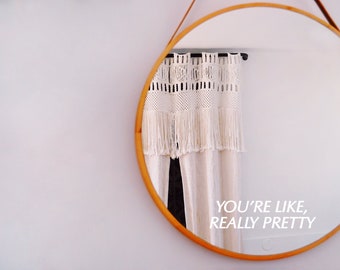Op de afbeelding: Een ronde spiegel met een lichthouten frame die een wit macramé gordijn met franjes reflecteert, met de tekst "YOU'RE LIKE, REALLY PRETTY" in het wit op het glazen oppervlak van de spiegel.