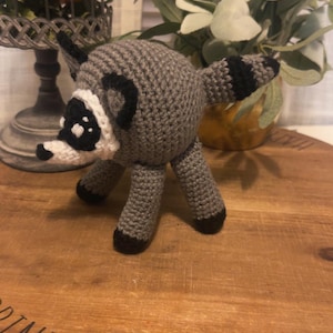 May include: A round, crocheted raccoon figurine featuring a gray body, a white face mask with black eye patches, black-tipped ears, and a striped tail. The raccoon has four gray legs with black feet and a small black nose, crafted with yarn.