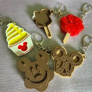 Puede incluir: Cinco llaveros con varios diseños: una copa de helado suave amarilla con un icono rojo de Mickey Mouse, un helado de palo marrón con forma de Mickey, un helado de palo rojo con forma de huella, una cara de Mickey marrón y un pretzel marrón.