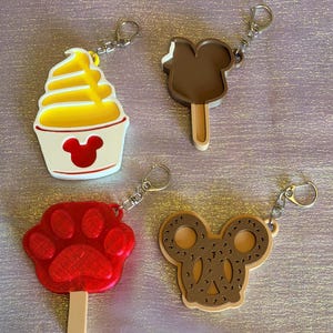 May include: Four keychains with silver rings. One is a yellow and white soft-serve ice cream with a red Mickey Mouse head. Another is a brown popsicle with a bite taken out. A red paw print and a Mickey Mouse pretzel complete the set.