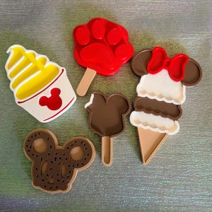 Puede incluir: Una colección de cortadores de galletas coloridos con forma de helado, una huella de pata y un pretzel de Mickey Mouse. Los diseños de helado incluyen un helado suave amarillo, un helado de chocolate y un cono de helado de Minnie Mouse con un lazo rojo.