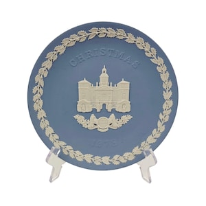 Puede incluir: Un plato redondo de cerámica azul con decoraciones en relieve de color blanco. El centro muestra un edificio, la palabra CHRISTMAS y el año 1978. Un borde de hojas de acebo blancas rodea el borde. El plato se apoya en un soporte de plástico.