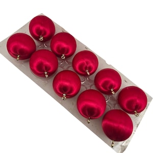 May include: A set of ten spherical red ornaments with a smooth satin finish, each featuring a small gold-toned metal loop at the top for hanging. The ornaments are arranged in a clear plastic tray with individual molded compartments.