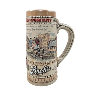 May include: A beige ceramic beer stein with a detailed village scene and the text "Stroh's". The stein has a large handle and a decorative rim. The text "Peter Stroh served guests at his legendary fire-brewed beer" is also visible.