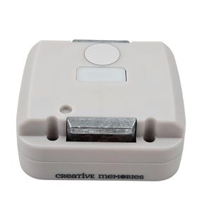 Könnte beinhalten: Ein hellgraues Creative Memories Papierstanzwerkzeug. Das Werkzeug hat eine rechteckige Form mit einer Metallklinge und dem Schriftzug "creative memories" auf der Unterseite.