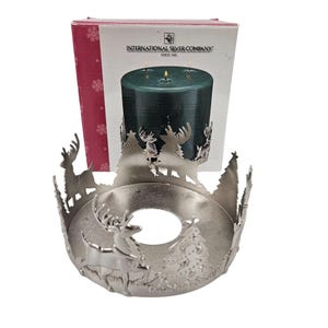 Puede incluir: Un portavelas de metal plateado con temática navideña. El soporte presenta diseños recortados de ciervos y pinos alrededor del borde. Una vela verde oscuro está dentro, con la caja de International Silver Company en el fondo.