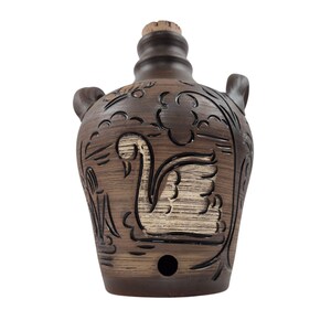 May include: A brown ceramic jug with a swan design. The jug has a cork stopper and two small handles. The swan is depicted in a pond with decorative foliage. The jug is approximately 8 inches tall.