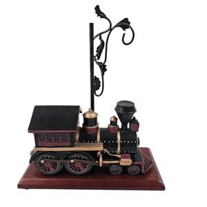 Puede incluir: Una figura decorativa de locomotora negra y burdeos con detalles dorados, montada sobre una base de madera oscura. Un elemento decorativo de metal negro con diseños en forma de hojas se extiende hacia arriba.
