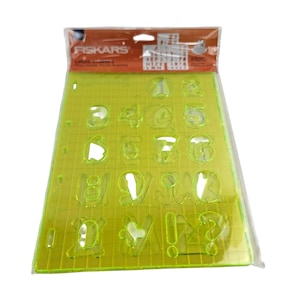 Könnte beinhalten: Eine Fiskars-Schablone in einer transparenten Kunststoffverpackung. Die Schablone hat eine transparente chartreuse Farbe und zeigt Zahlen, Buchstaben und Symbole. Das Fiskars-Logo ist oben auf der Verpackung zu sehen.