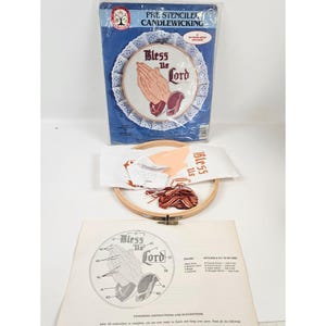 May include: A pre-stenciled candlewicking kit featuring a design of praying hands with the words "Bless Us Lord." The kit includes a hoop, fabric, thread, and instructions. The design is framed by a white lace trim.