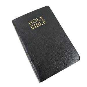 Puede incluir: Una Biblia negra de tapa dura con letras doradas que dicen "HOLY BIBLE". El libro tiene una cubierta texturizada y bordes de página dorados. El libro está ligeramente inclinado, mostrando el lomo y la portada.