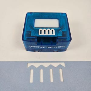 Puede incluir: Un troquelador de papel azul Creative Memories con un diseño blanco y el texto "Creative Memories". Debajo hay un papel azul claro con un diseño recortado a juego. Suministros para álbumes de recortes.