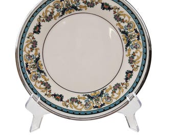 May include: A white ceramic plate with a silver rim and a decorative border featuring teal, yellow, and purple floral patterns. The plate is displayed on a clear plastic stand, showcasing its design. This elegant plate is suitable for serving food.