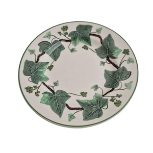 May include: A round, off-white plate with a green border. The plate is decorated with a green ivy leaf and vine pattern. The leaves are detailed with shading, and the vines are brown. A classic design for serving food.