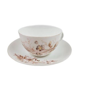 May include: A white teacup and saucer set with a floral design in brown and gray. The teacup has a gold rim and a handle. The saucer has a matching floral pattern. The set is on a white background.