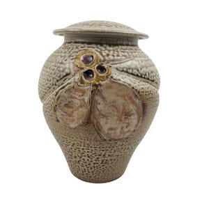 May include: A ceramic urn with a textured, speckled surface in shades of tan and brown. The urn features a lid and is adorned with a decorative cluster of three dark purple orbs and two brown, textured, organic shapes.