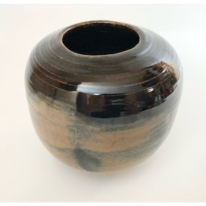 May include: A round, brown ceramic vase with a glossy finish. The vase has a wide opening and a slightly rounded bottom. The glaze is a dark brown with hints of gold and black.