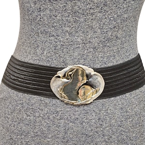May include: Black elastic belt with a silver and gold oval buckle. The buckle features a sculpted design with a central gold element and silver framing. The belt is worn over a gray dress.
