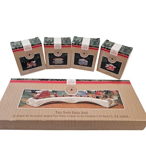 May include: A train trestle display stand with four boxed train ornaments. The boxes are brown with green and red accents. The display stand is beige with a train scene. Text on the box reads "Keepsake Ornament".